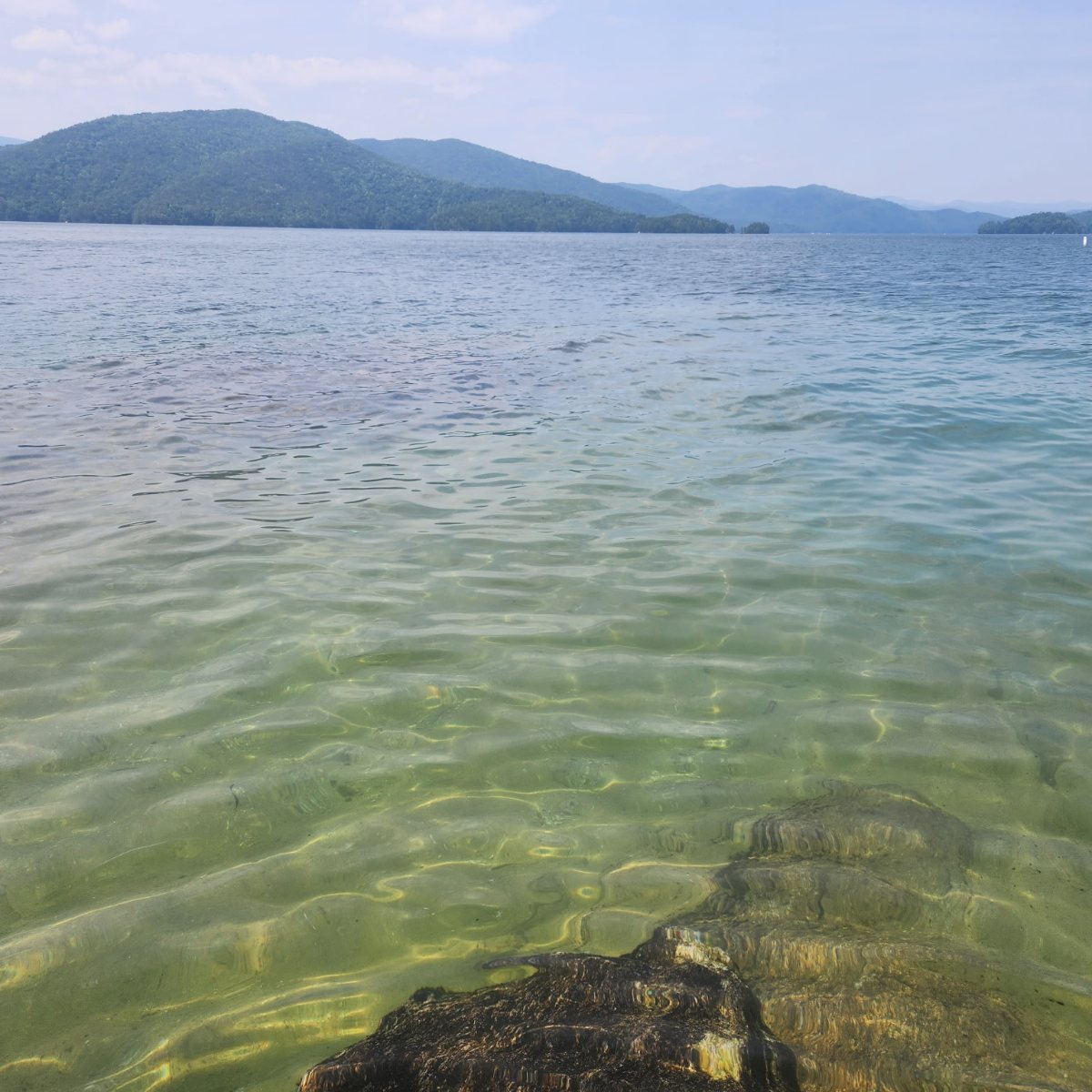 Clear green water over a rock at the shoreline, looking out across Lake Jocassee toward forested mountains.