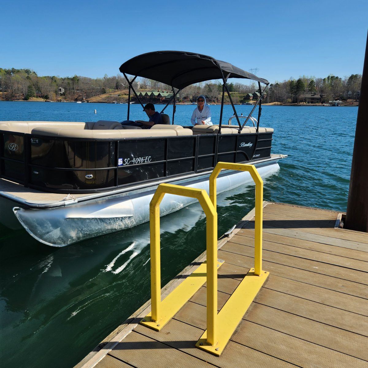 A black tritoon with tan seating easing up to a floating dock with a yellow boarding rail on a clear spring day.